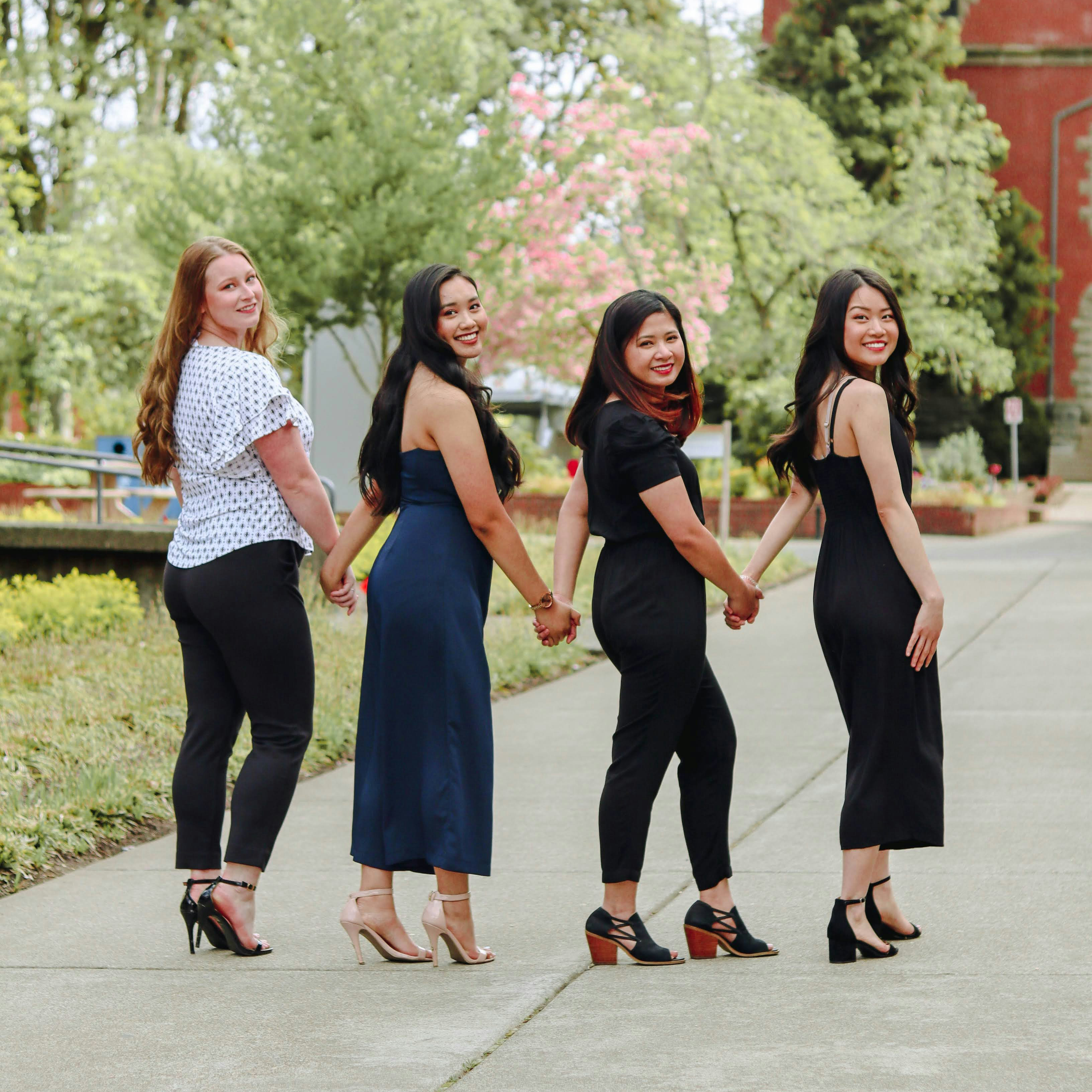  four girls in a group holding hands on a sidewalk in front of trees and a red building