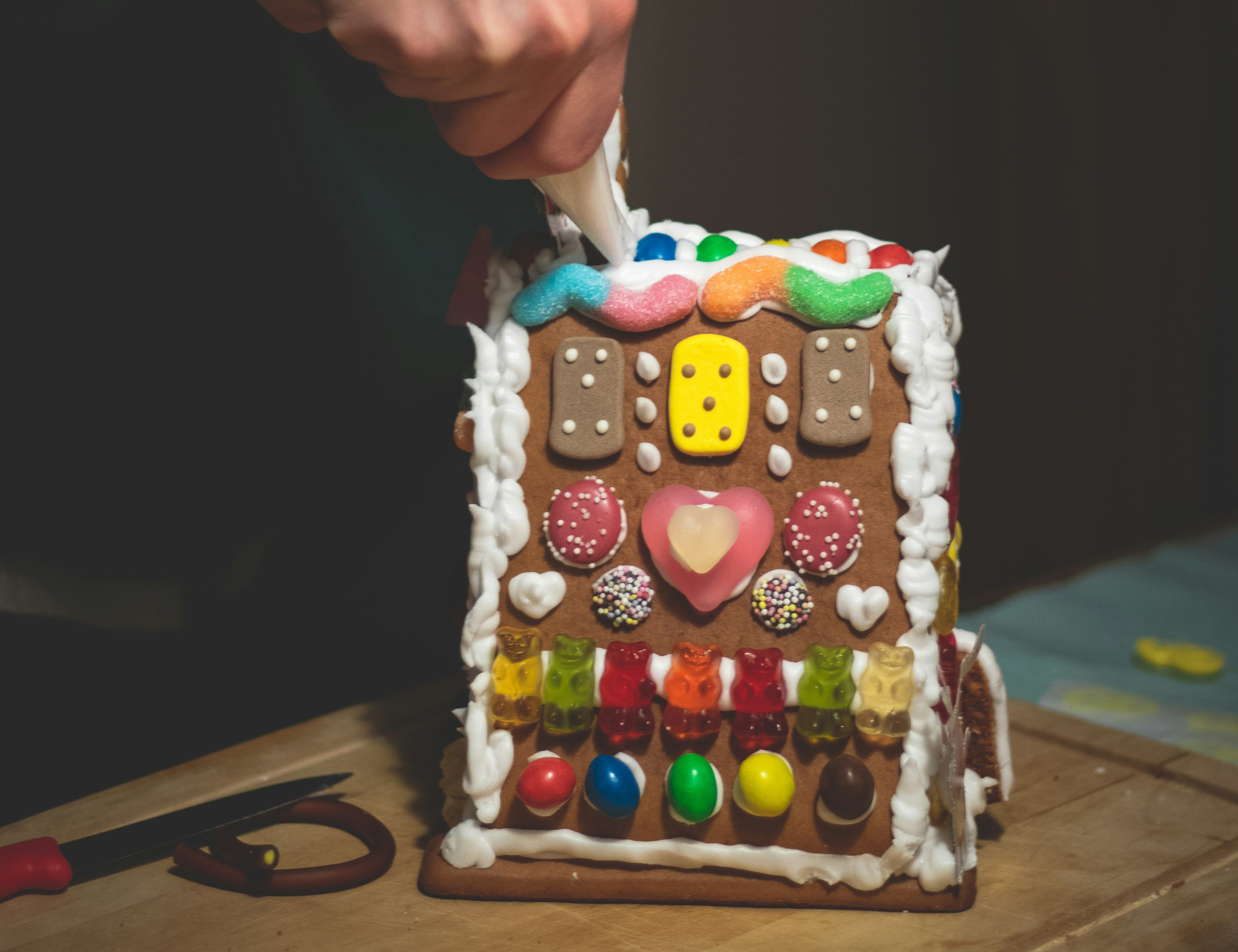 a person decorating a gingerbread house with colourful icing and candy decorations by hand on a wooden countertop